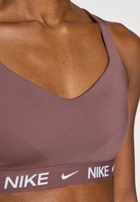 Καφέ αθλητικό σουτιέν από απαλό, ελαστικό ύφασμα. Διαθέτει V-λαιμόκοψη και λευκό λογότυπο Nike στη ελαστική ταινία.