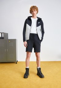 Zip-up hoodie i sort og grå, hvid t-shirt, sorte shorts, sorte snørebåndsstøvler. Struktureret stof, sporty design og synlige logo-detaljer.