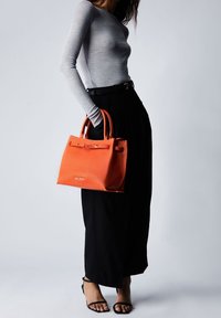 Sac en cuir orange texturé avec poignées doubles et un fermoir à l'avant, associé à un haut gris ajusté à manches longues et un pantalon noir à pattes larges.