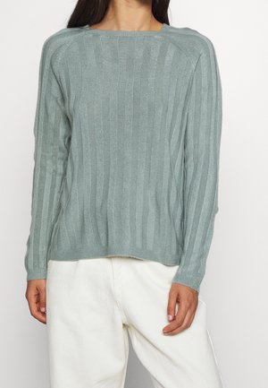 Vrouw draagt een lichtgrijze ribgebreide trui met lange mouwen en losse witte broek, staand tegen een effen witte achtergrond.