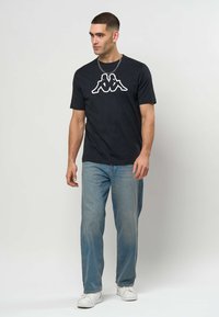 Čierne bavlnené tričko s bielym logom uprostred, doplnené svetlomodrými džínsami a bielymi teniskami. Jednoduché, ležérne oblečenie.