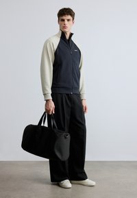 Laivastonsininen ja kermanvärinen vetoketjulla varustettu takki, jossa on korkea kaulus, yhdistetty mustiin housuihin. Malli pitää kädessään mustaa duffel-laukkua ja käyttää valkoisia tennareita.