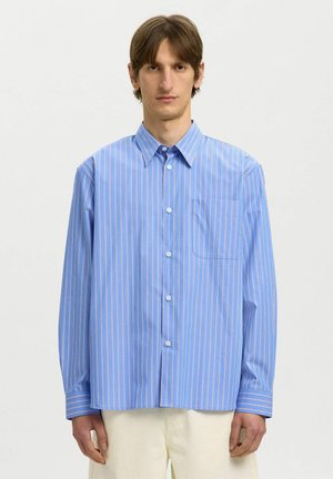 Homme portant une chemise oversize bleu clair à fines rayures verticales roses et blanches, et un pantalon écru, debout devant un fond uni.