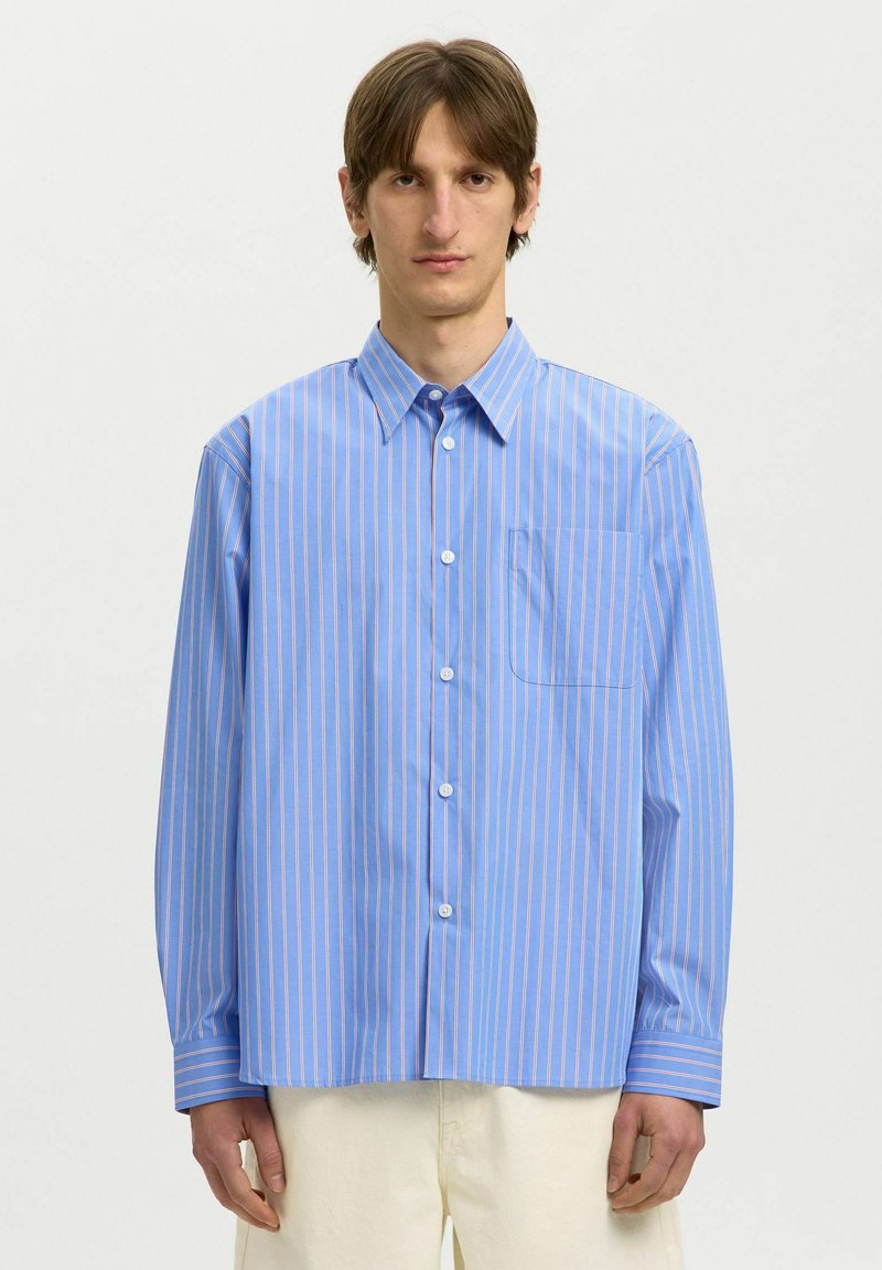 Homme portant une chemise oversize bleu clair à fines rayures verticales roses et blanches, et un pantalon écru, debout devant un fond uni.