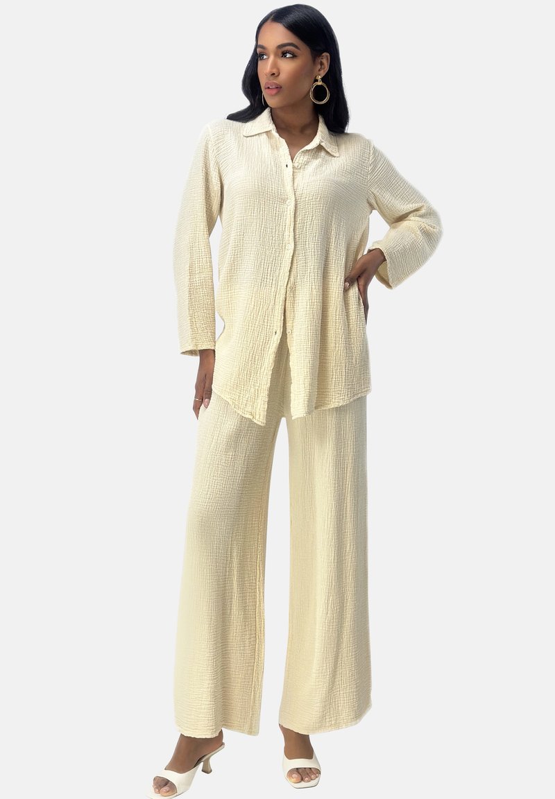 Completo due pezzi color crema composto da una camicia con bottoni in tessuto testurizzato e pantaloni a gamba larga. Accentuato da sandali con tacco.