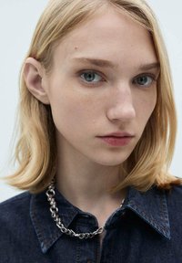 Cheveux blonds aux ondulations douces, peau claire, portant une chemise en denim avec un collier en chaîne. L'arrière-plan neutre met en valeur les traits du visage.