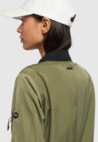 Giubbotto bomber verde oliva con colletto a costine nero, tasca laterale con zip e texture liscia. Berretto bianco parzialmente visibile sopra.