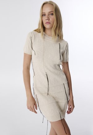 Beige gebreide jurk met korte mouwen, versierd met contrasterende zwarte losse draadjes langs de naden en een ribbelboord, met een aansluitend ontwerp.