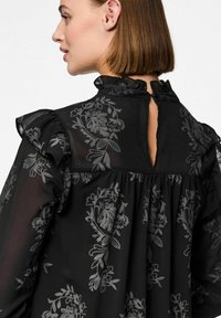 Černá blůza s květinovým vzorem z průsvitné látky, volánkovým límcem, otvorem na zádech a nařaseným pasem. Obsahuje zapínání na knoflík u krku.