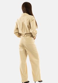 Mono utilitario beige con cuello tipo collar, cintura elástica y diseño holgado. Presenta bolsillos traseros y una textura de tela suave.