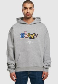 Sudadera gris con un bolsillo delantero, que presenta un gráfico colorido de "BKLYN" y el texto "Spread Love It's the Brooklyn Way" debajo. Material suave.