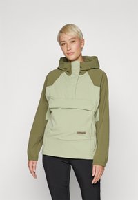 Lysegrønn anorakkjakke med olivengrønn raglanermer, justerbar hette, frontlomme og logoaksent. Materialet ser glatt og lett ut.