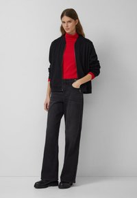 Eine Frau trägt eine schwarze Jacke mit Reißverschluss, einen roten Rollkragenpullover, weite schwarze Jeans und schwarze Stiefel und steht vor einer schlichten hellgrauen Wand.