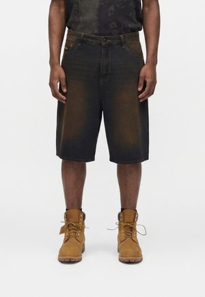 Mand iført sorte denimshorts og brune arbejdstøvler, stående mod en ensfarvet hvid baggrund.