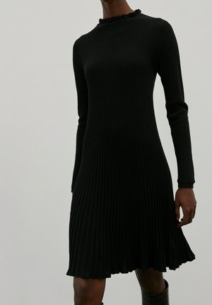 Femme portant une robe en maille côtelée noire à manches longues avec un col et un ourlet à volants, associée à des bottes noires.