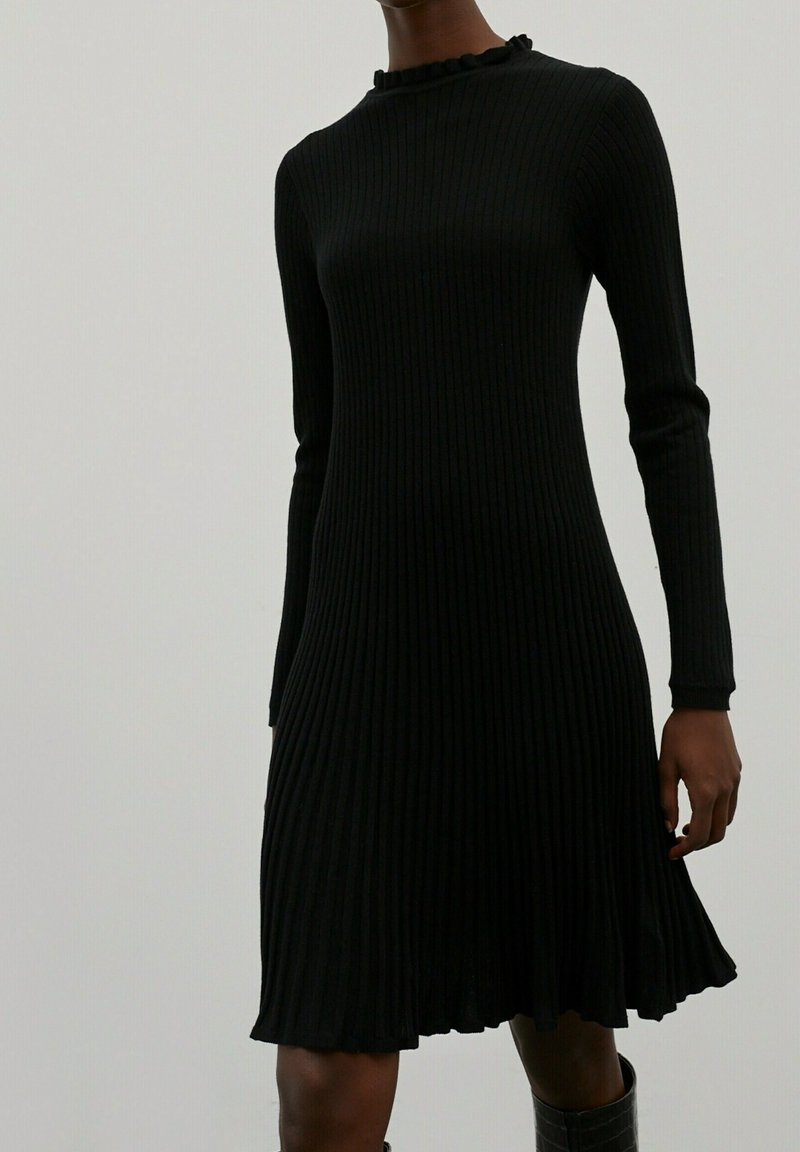 Femme portant une robe en maille côtelée noire à manches longues avec un col et un ourlet à volants, associée à des bottes noires.