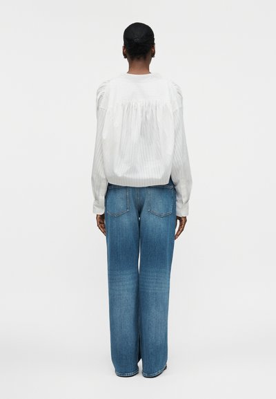 Άτομο με σκουρόχρωμο δέρμα και μαλλιά δεμένα πίσω στέκεται με την πλάτη στραμμένη, φοράει λευκό πουκάμισο μακρυμάνικο και μπλε τζιν παντελόνι με φαρδιά πόδια, σε απλό φόντο.