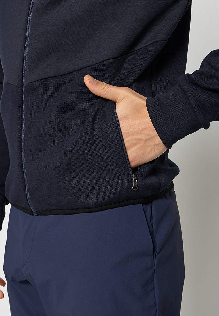 Giacca zip-up blu navy realizzata in tessuto testurizzato, con tasca laterale con zip e orlo nero a contrasto. Indossata con pantaloni blu scuro, evidenziando la posizione delle mani.