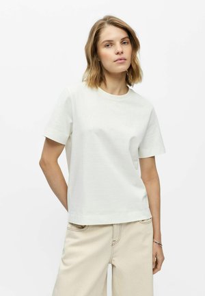 Vrouw met schouderlang blond haar, draag een effen wit T-shirt met korte mouwen en beige broek, staand voor een witte achtergrond.