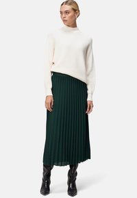 Pull ivoire à côtes avec col haut associé à une jupe midi plissée verte ; style avec des bottes noires montantes. Texture lisse et forme en A.