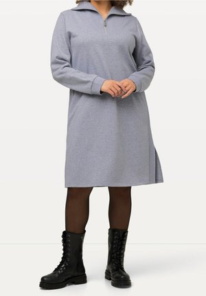Femme portant une robe à manches longues gris clair avec un col zippé, des collants noirs transparents et des bottes de combat noires à lacets, debout les mains jointes.