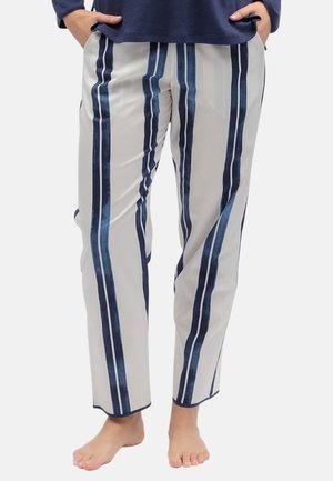 Persona con pantaloni bianchi con strisce verticali blu scuro, in piedi a piedi nudi con le mani in tasca davanti a uno sfondo semplice.