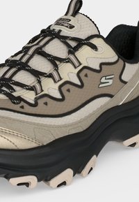 Baskets Skechers beige et noires avec semelle noire épaisse, lacets noirs et beiges, et tige en tissu mesh avec logo de la marque sur le côté.