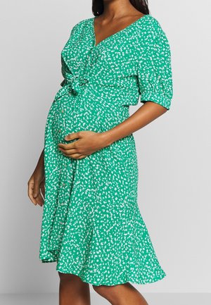 Femme enceinte portant une robe verte avec un imprimé floral blanc, tenant son ventre d'une main, sur un fond gris uni.
