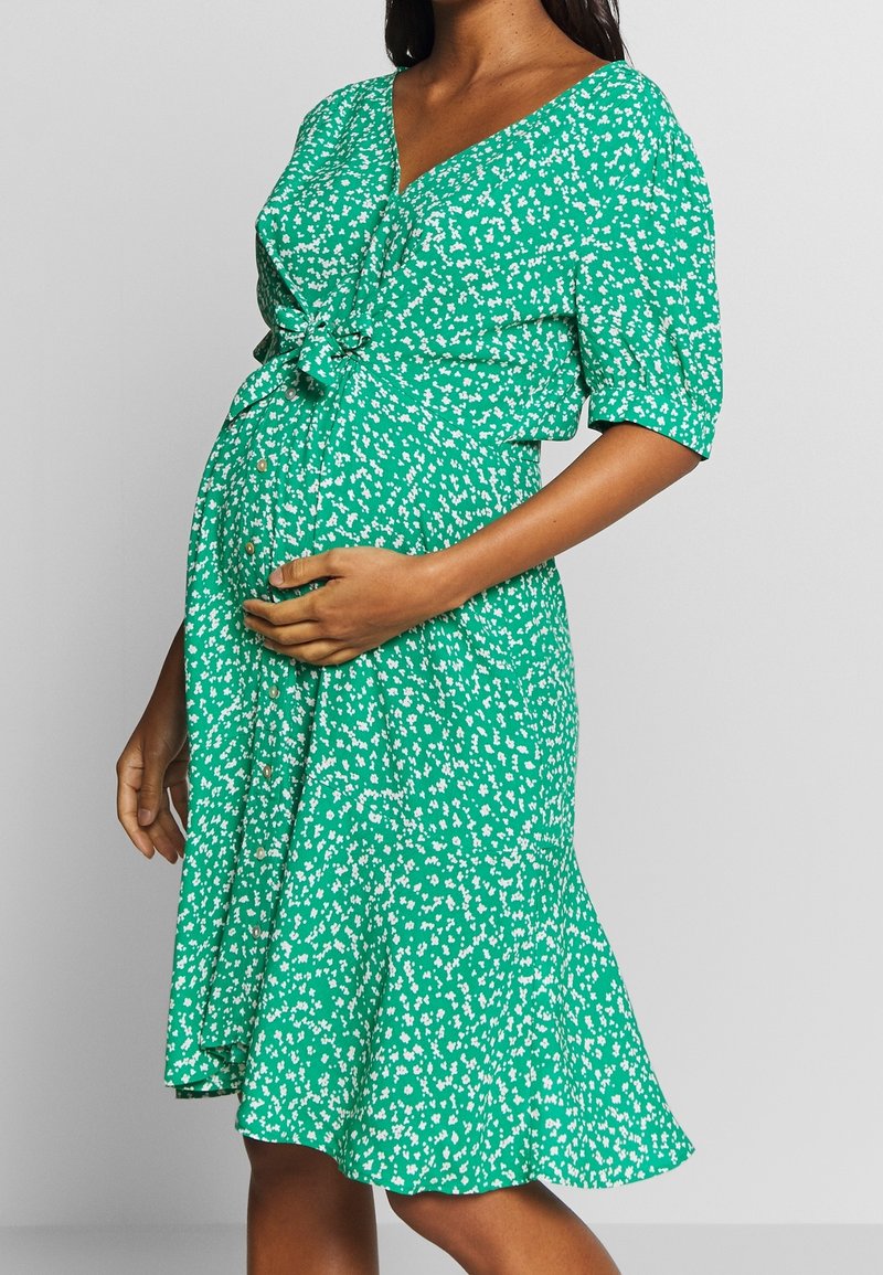 Femme enceinte portant une robe verte avec un imprimé floral blanc, tenant son ventre d'une main, sur un fond gris uni.