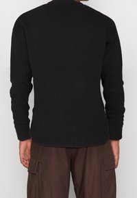 Pull noir en tricot avec poignets et ourlet côtelés. Doté d'un col rond et d'une texture lisse. Porté avec un pantalon cargo marron.