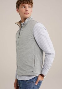Pelēks bezpiedurkņu veste ar augstu apkakli un rāvējslēdzēju, teksturēta auduma, uzvilkta virs gaiši zila un balta svītrainā garajiem piedurknēm krekla.