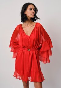 Rotes transparentes Kleid mit ausgestellten Ärmeln, tiefem V-Ausschnitt und einem rüschierten Saum. Es hat eine taillierte Taille mit einer dekorativen Gürtelschnalle.