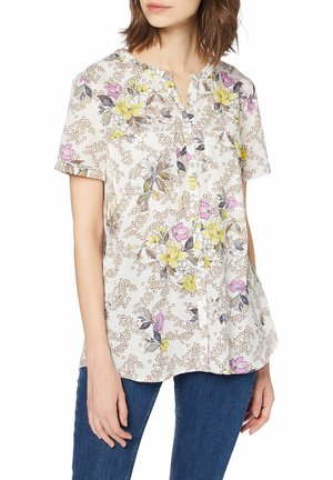 Femme portant un chemisier à manches courtes blanc avec des motifs floraux jaunes et roses ainsi que des points circulaires, associé à un jean en denim bleu.