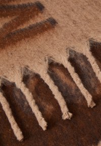 Tissu tissé marron et beige avec une texture lisse, comportant des franges claires sur un bord et des motifs abstraits sombres sur toute la surface.