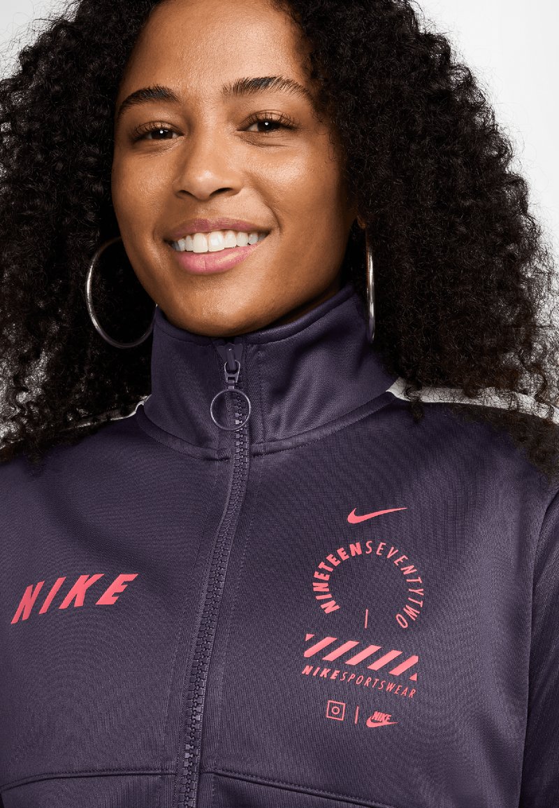 Chaqueta con cremallera de Nike en púrpura oscuro, con un logo redondo y texto en rosa. Textura suave con herrajes plateados en el cuello y los bolsillos.