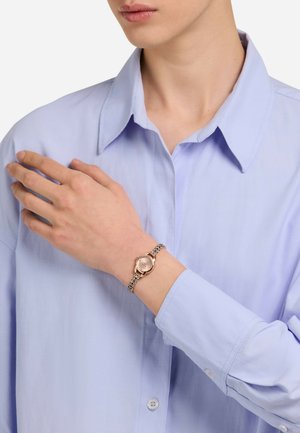 Różowo-złoty zegarek z okrągłą tarczą i bransoletą z ogniwami w kolorze srebra, noszony na ręce na tle jasnoniebieskiej koszuli z kołnierzykiem.