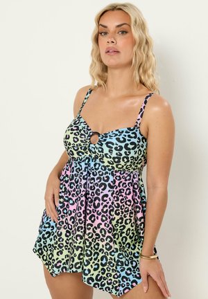 Vrouw met golvend blond haar die een mouwloze, meerkleurige jurk met luipaardprint draagt met een ringendetail op de borst, staand tegen een witte achtergrond.