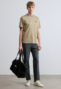 Bézs pamut póló kerek nyakkivágással és apró logóval, szürke szabott nadrággal és fehér tornacipővel párosítva. Fekete táska logóval a kézben.