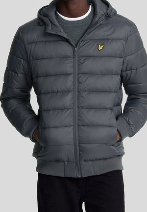 Homme portant une doudoune matelassée gris foncé avec capuche, fermeture éclair à l'avant, poignets côtelés et un petit logo d'oiseau jaune sur la poitrine.