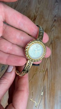 Χέρι που κρατά ένα χρυσό ρολόι χειρός Versace με μοτίβο στο λουράκι και στρογγυλό καντράν διακοσμημένο με στρας, δείχνοντας 3:15.