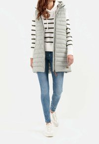 Lichtgrijze gewatteerde bodywarmer met capuchon, ritssluiting en zijzakken. Draagt over een witte gestreepte hoodie en blauwe jeans, gecombineerd met witte sneakers.