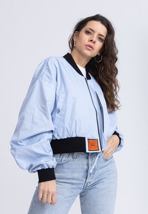 Hellblaue Bomberjacke mit schwarzen, gerippten Bündchen und Kragen. Verfügt über einen Reißverschluss vorne, eine Seitentasche und ein oranges Etikett am Saum.