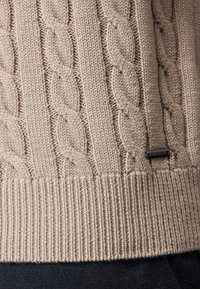 Beiger gestrickter Pullover mit Zopfmuster, geripptem Saum und dezentem Logotag. Weiche Textur mit sichtbaren Stichen und dickem Garn.