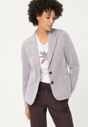 Woman with curly hair wearing a striped blazer over a white t-shirt with a palm tree design and dark drawstring pants.