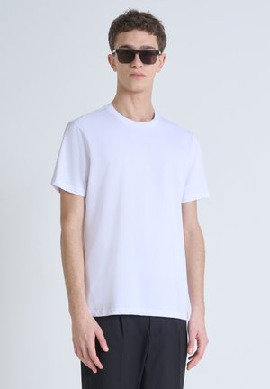 Camiseta blanca de manga corta con cuello redondo, hecha de una tela suave; combinada con pantalones negros y gafas de sol oscuras.