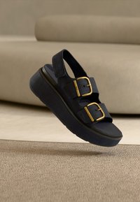 Μαύρο πλατφόρμα σανδάλι με δύο φαρδιές λουριά που διαθέτουν χρυσά αγκράφες, τοποθετημένο πάνω σε μπεζ χαλί με φόντο καναπέ του ίδιου χρώματος.