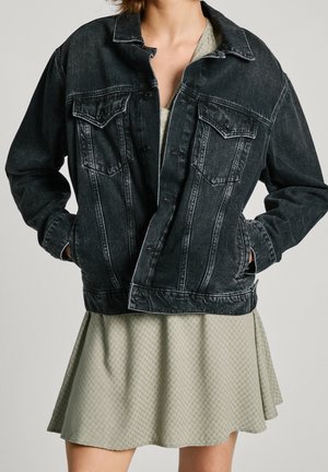 Schwarze Jeansjacke mit Knopfverschluss, zwei Brusttaschen und einem leicht übergroßen Schnitt, kombiniert mit einem hellgrünen gemusterten Kleid.