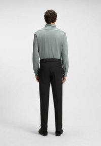 Ein Mann trägt ein hellgrünes langärmliges Hemd, das in schwarze formelle Hosen gesteckt ist, und steht mit dem Rücken zu einem schlichten weißen Hintergrund.