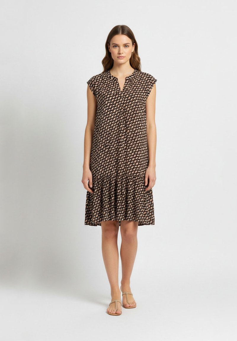 Femme debout portant une robe sans manches noire et beige à motifs, longueur genou, avec un ourlet à volants et des sandales beiges sur un fond uni.