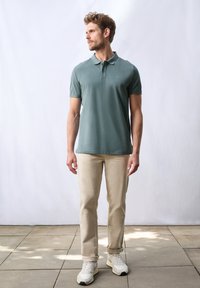 Mäns turkosa polo t-shirt med krage och korta ärmar, kombinerad med beiga byxor. Modellen står på stenplattor, iklädd ljusa sneakers.